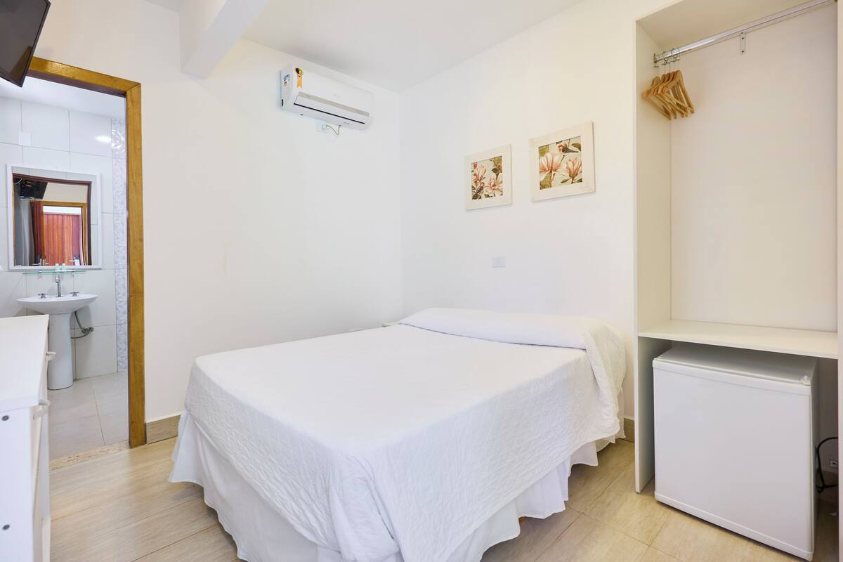 A clean, white room with a bed, a small white cabinet, a mirror, and a wall-mounted air conditioner.