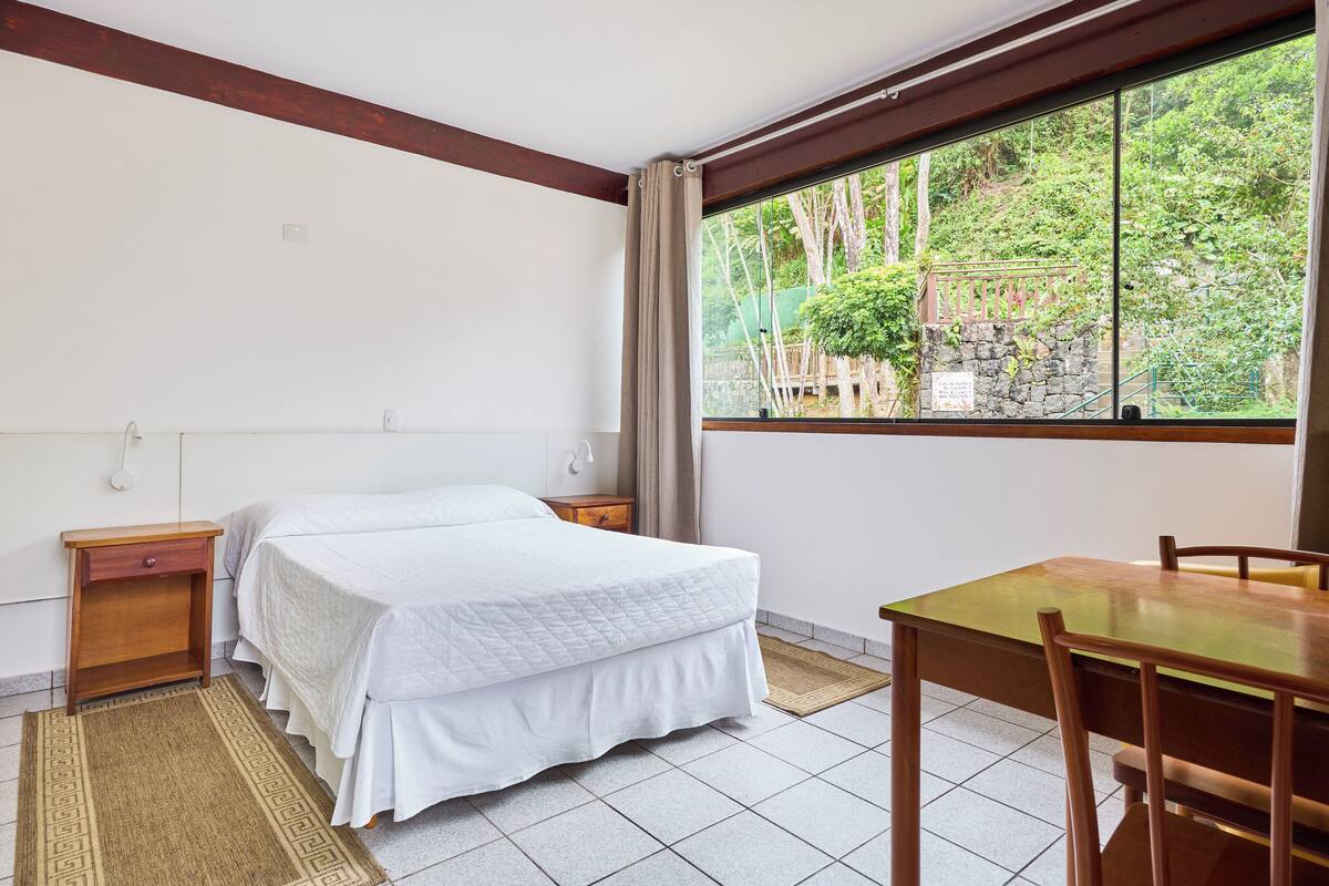 A bedroom with a bed, bedside tables, a wooden desk, and a window with a view of greenery.