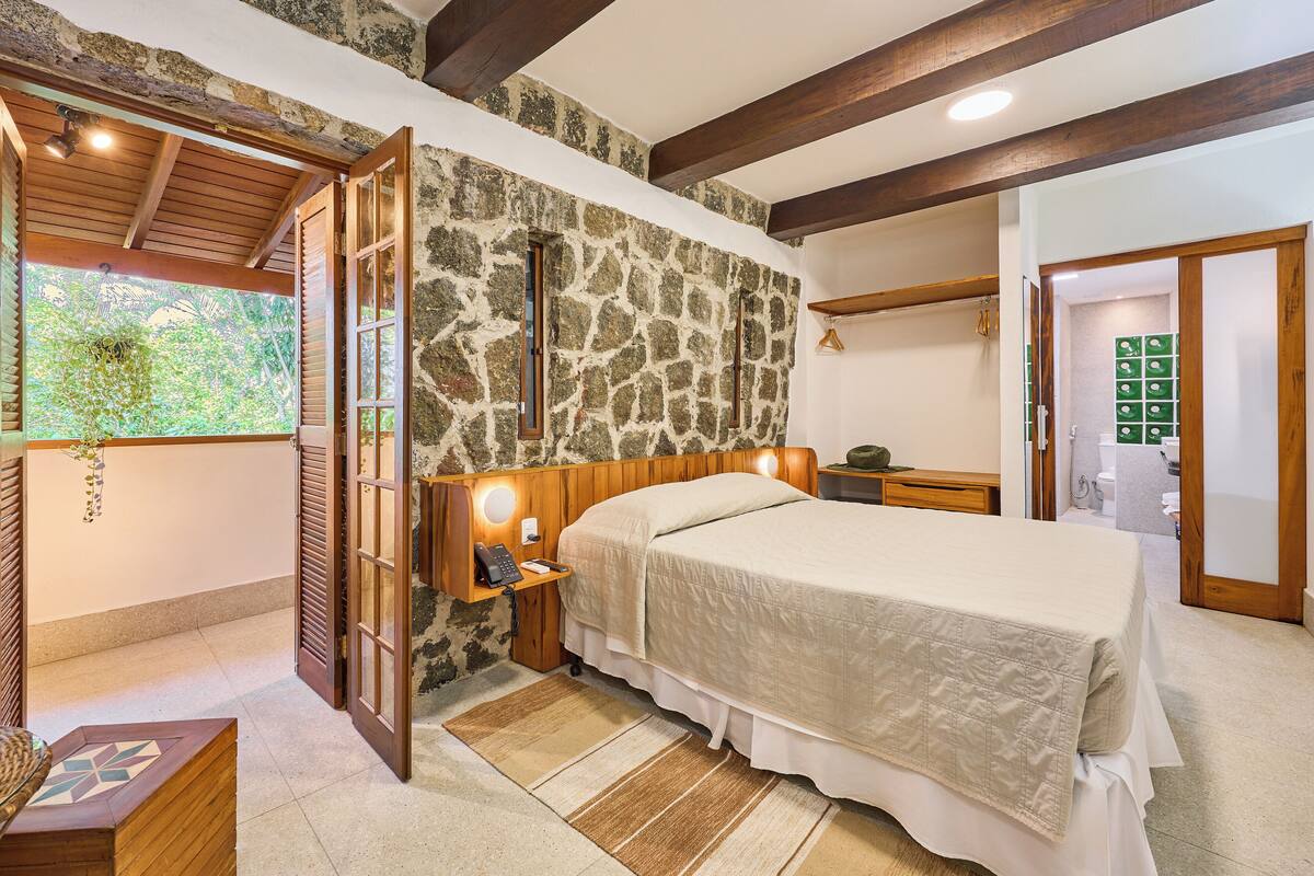A bedroom with a stone wall, wooden ceiling beams, a bed, bedside table, and a window with a view of greenery.