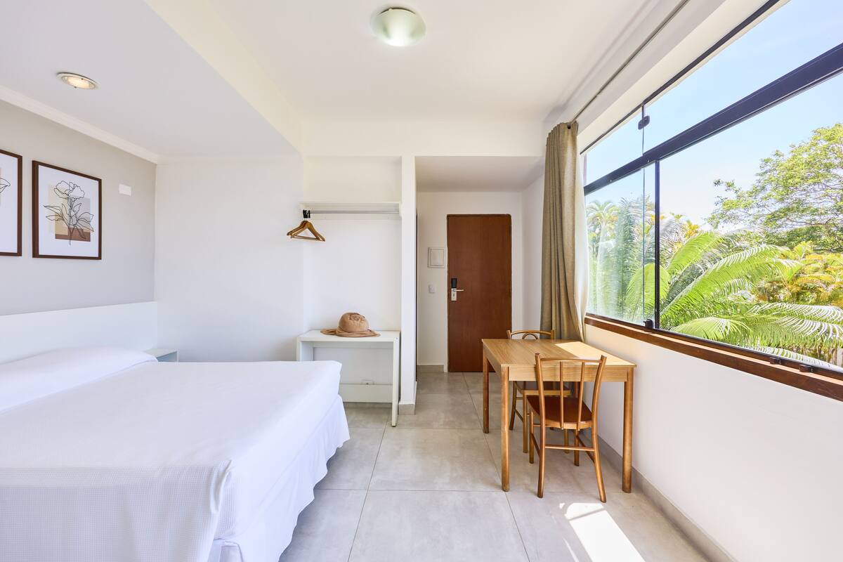 A hotel room with a bed, a small table, a chair, and a window with a view of greenery.