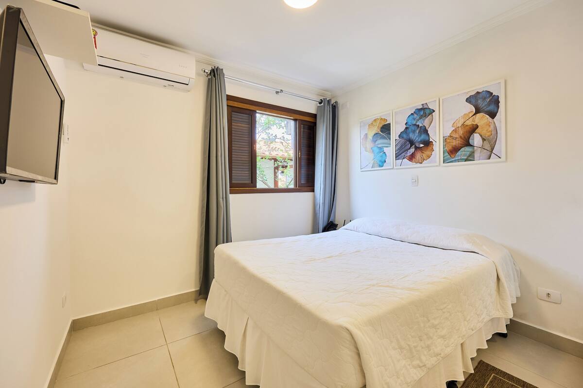 A bedroom with a bed, a window with curtains, an air conditioning unit, and a wall-mounted TV.