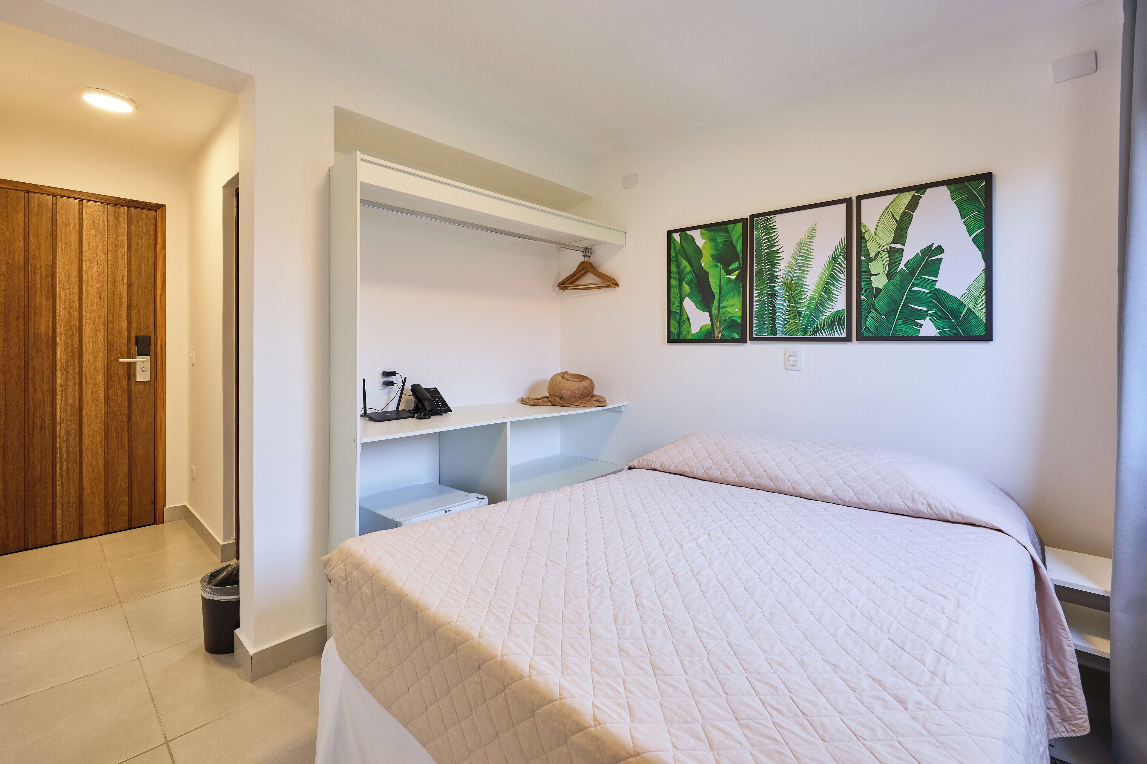 Un dormitorio con cama, una puerta de madera, pared blanca y repisa con sombrero y teléfono.
