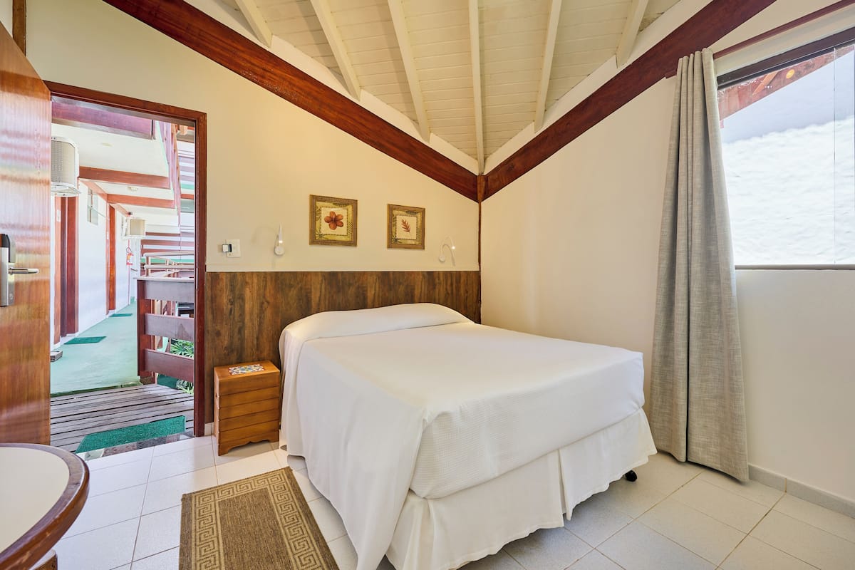 A bedroom with a bed, wooden ceiling beams, a small bedside table, and a window with curtains.