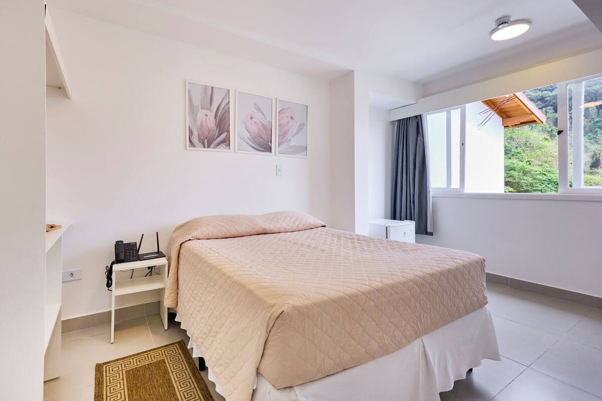 A bedroom with a bed, a bedside table, a window with a view of greenery, and three wall-mounted paintings.