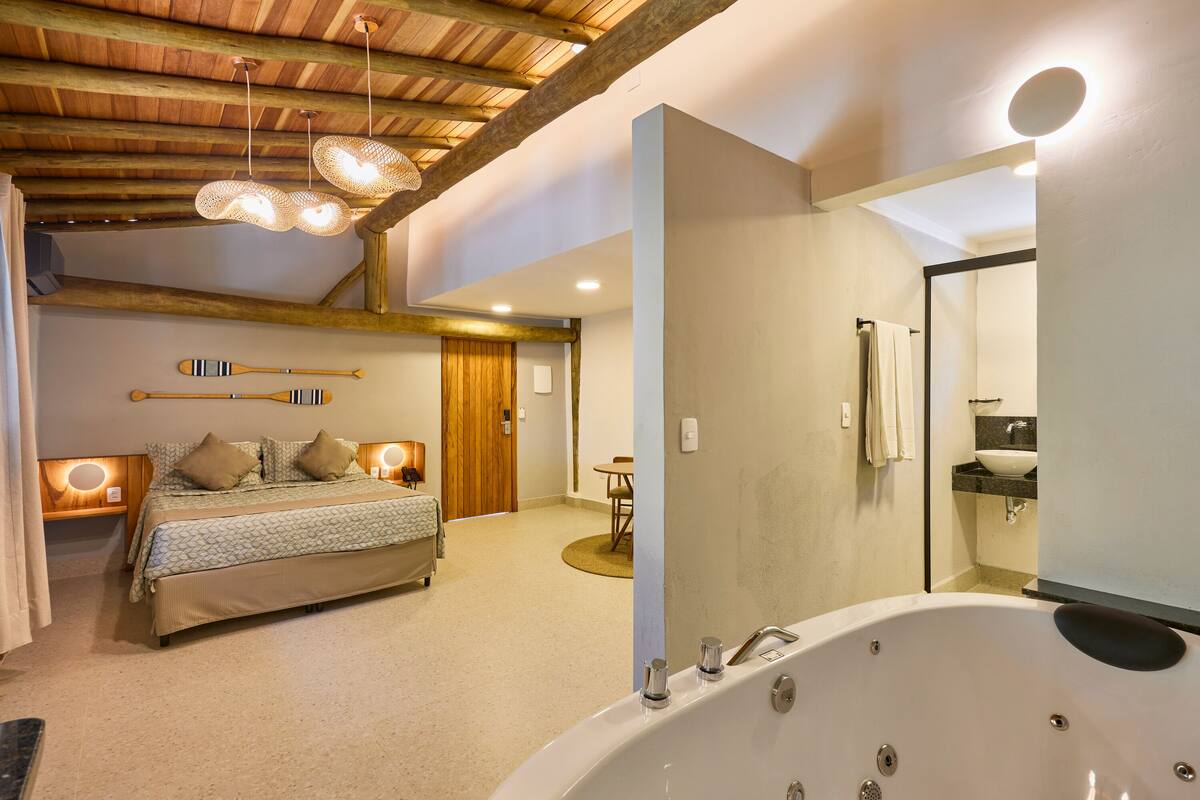 A bedroom with a large bed, a sitting area with a table and chairs, a bathroom with a bathtub, and a wooden ceiling.