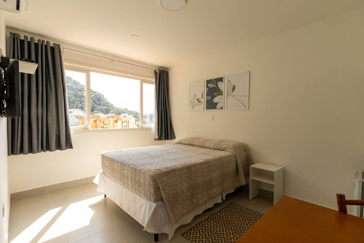 A bedroom with a bed, a small bedside table, a window with curtains, and a view of buildings.