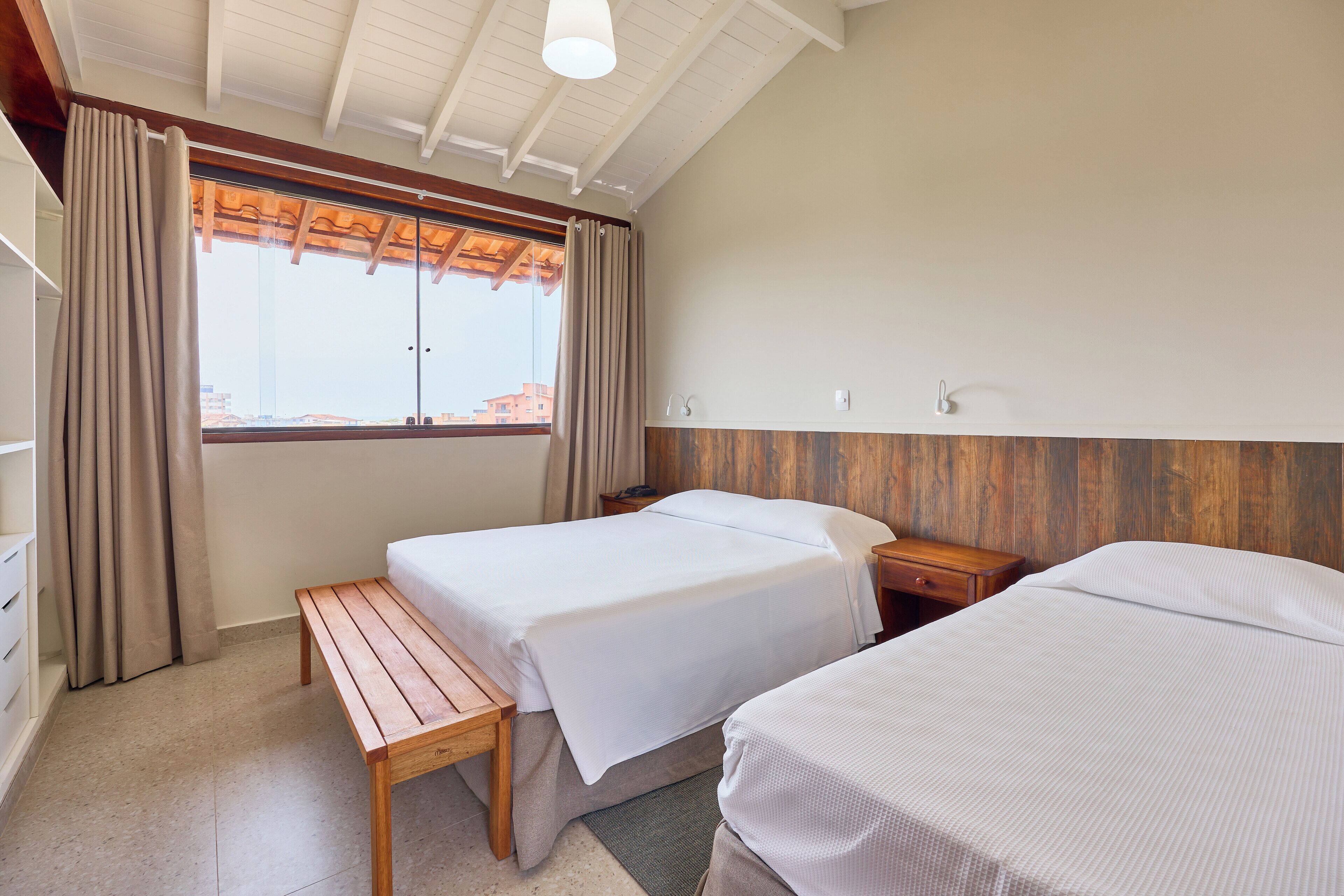 A hotel room with two beds, a wooden bench, a nightstand, and a window with curtains.