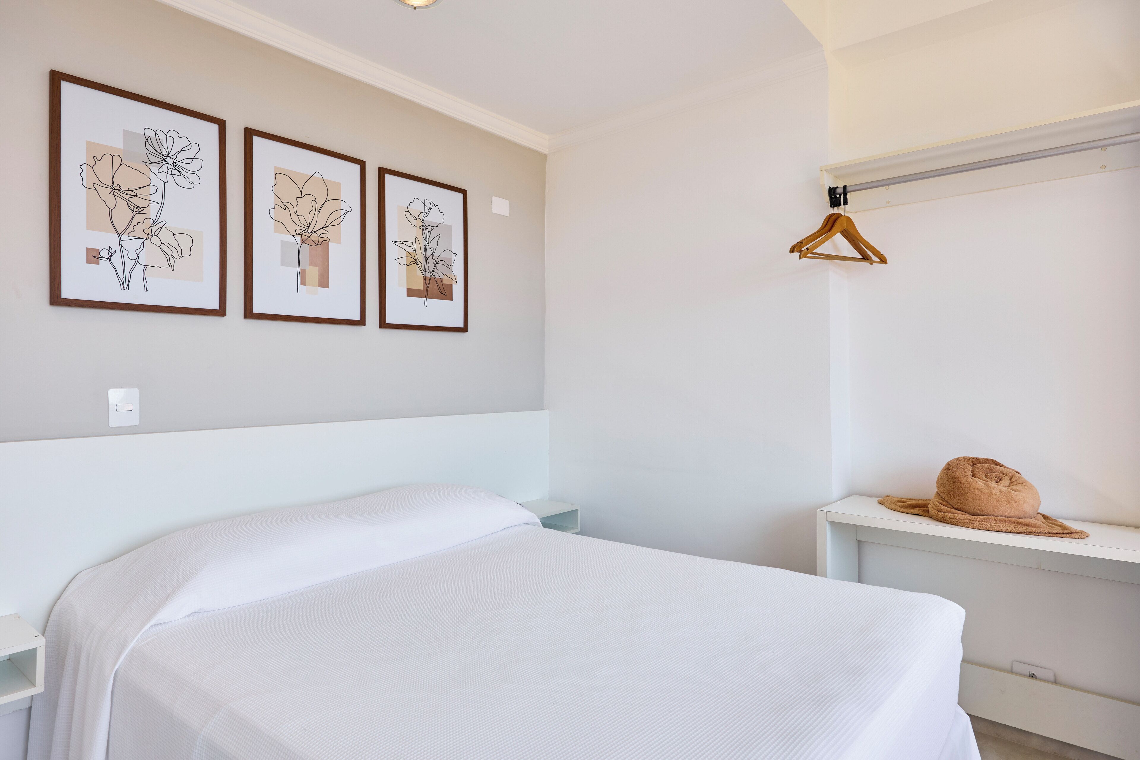 Un dormitorio con una cama, una mesita de noche, un perchero de madera colgante y tres cuadros con motivos florales en la pared.