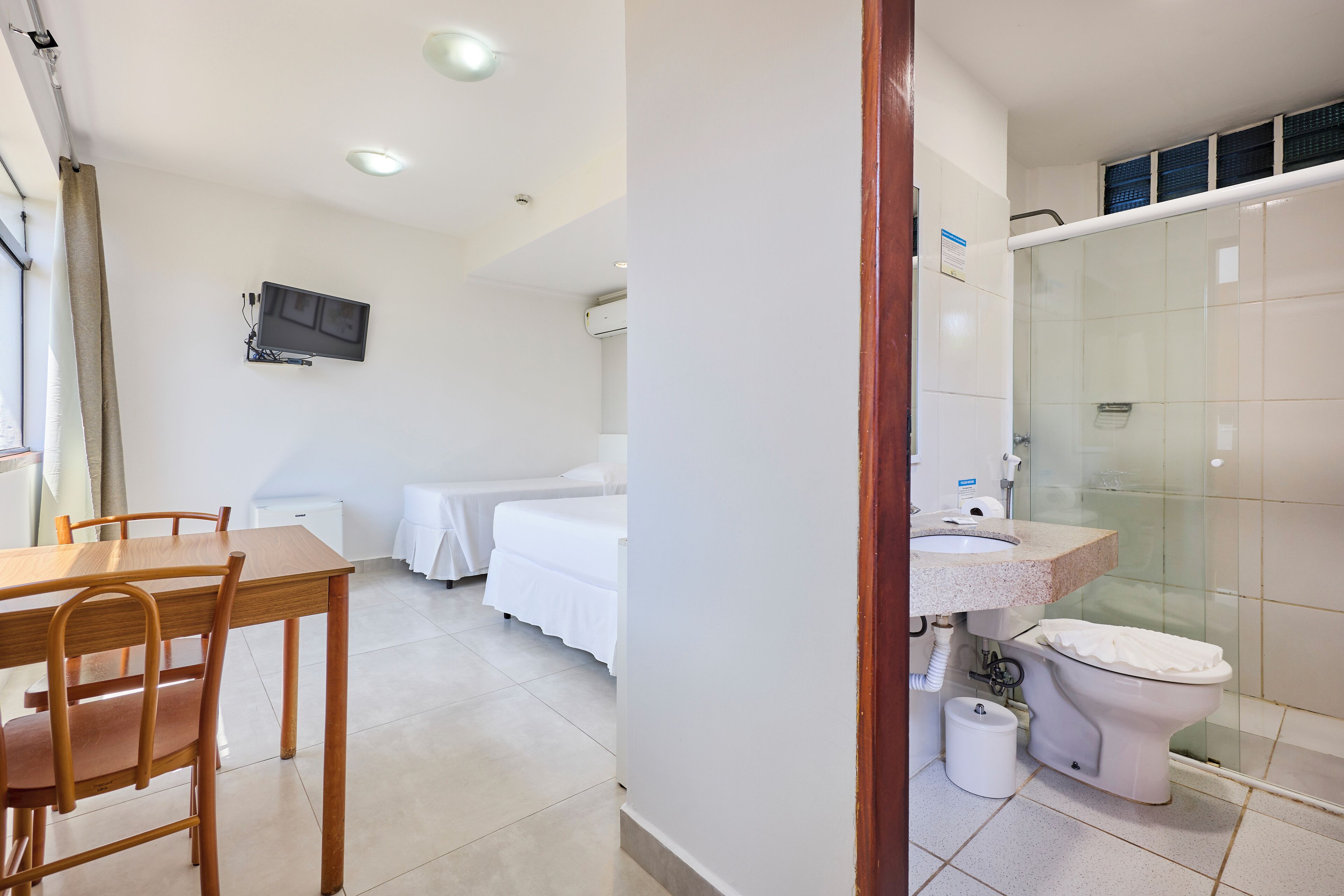 Habitación de hotel con una mesa de comedor de madera, una cama con ropa de cama blanca, un televisor montado en la pared y un baño con cabina de ducha de vidrio.