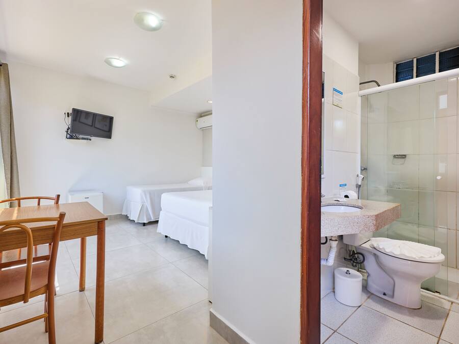 Habitación de hotel con una mesa de comedor de madera, una cama con ropa de cama blanca, un televisor montado en la pared y un baño con cabina de ducha de vidrio.