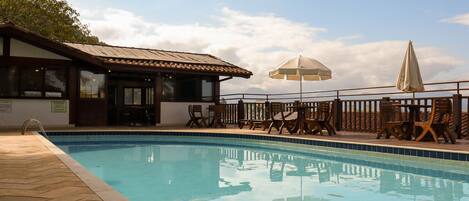 A swimming pool with wooden deck chairs and umbrellas, a building with a tiled roof, and a view of the sky.