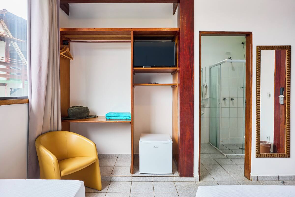 A compact room with a yellow chair, a white refrigerator, a wooden shelf, and a glass shower area.