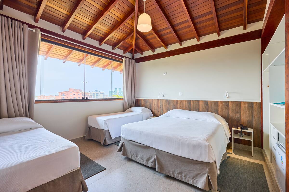 A hotel room with two beds, a wooden ceiling, a large window with curtains, and a small bedside table with a phone.