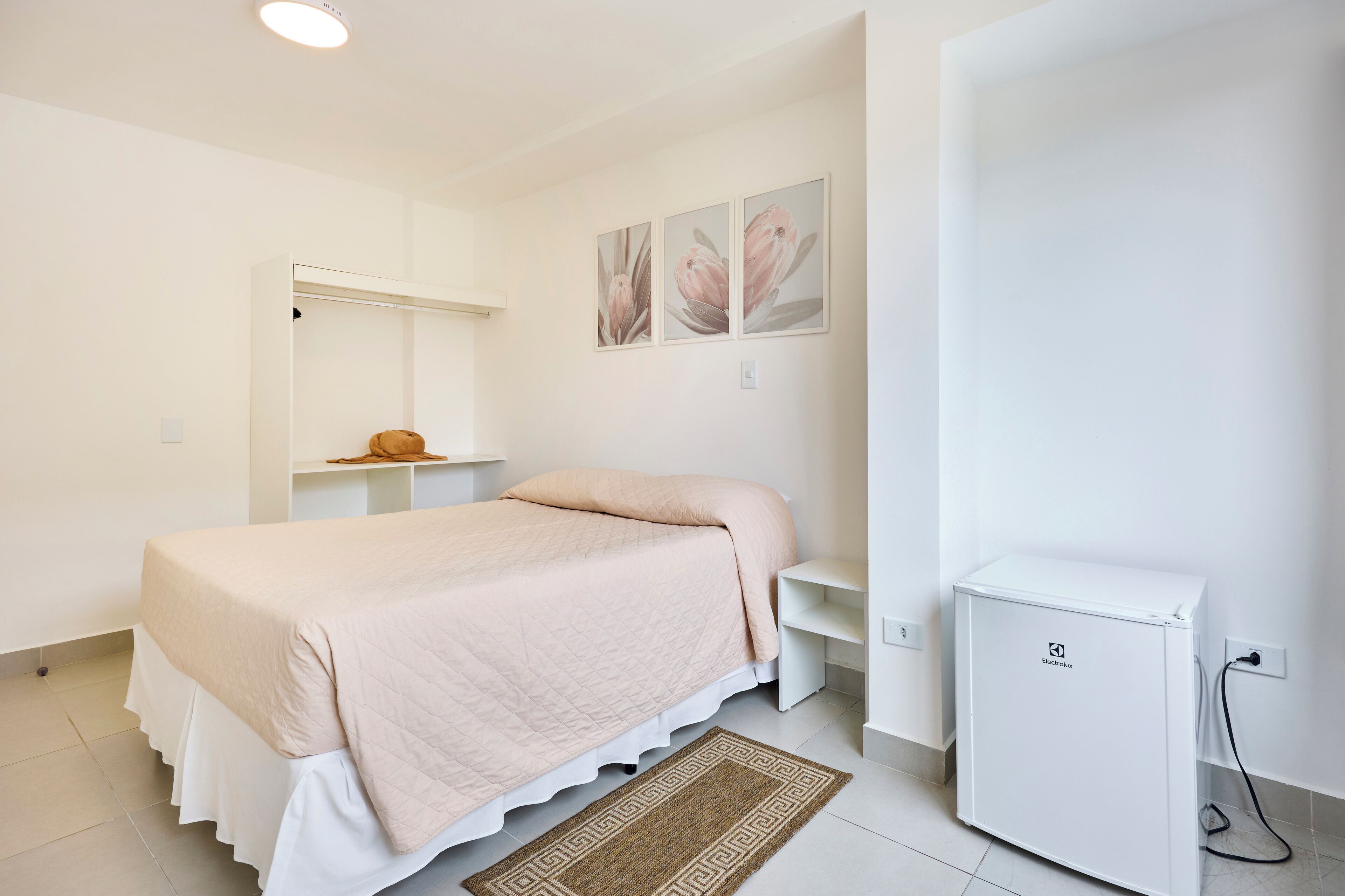 Une chambre comprenant un lit, une petite armoire blanche, une étagère fixée au mur et un réfrigérateur compact.