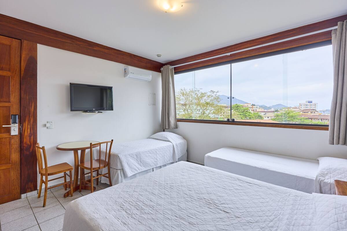 A hotel room with two beds, a TV, a small table with chairs, and a window with a view of buildings and trees.