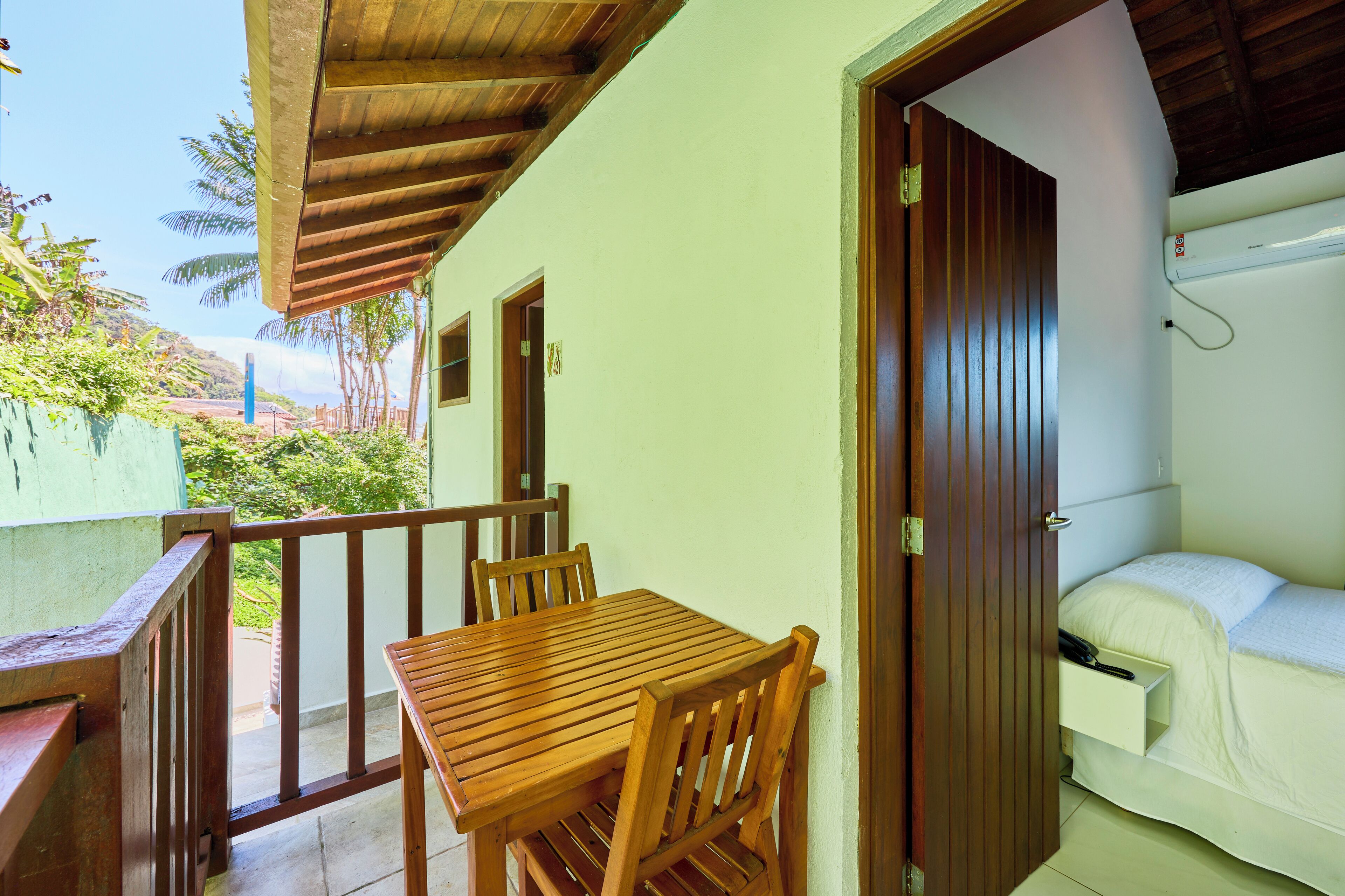 Quarto com mesa e cadeiras de madeira, cama com colchão branco e varanda com guarda-corpo de madeira.