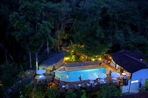 An outdoor swimming pool surrounded by wooden deck and lounge chairs, with a view of dense forest in the background.