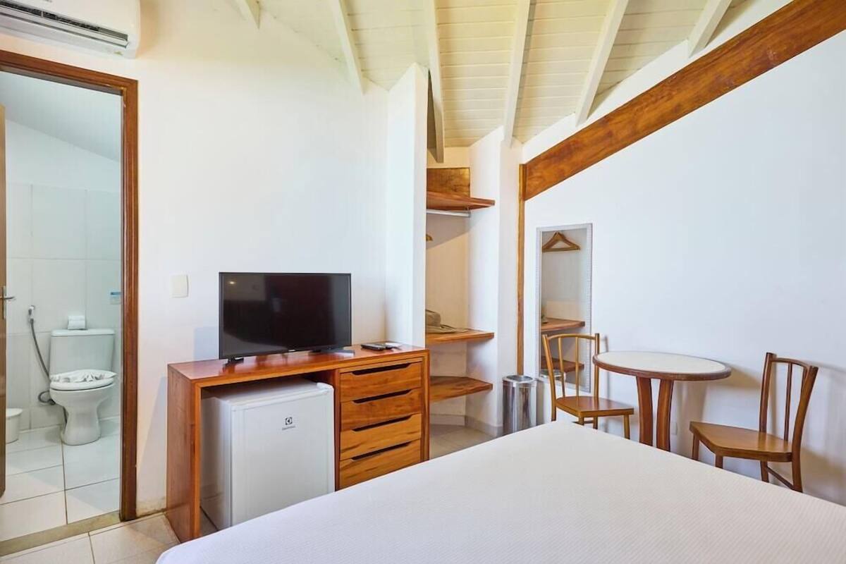 A bedroom with a bed, a TV on a wooden desk, a small fridge, a wooden chair, and a table with chairs.