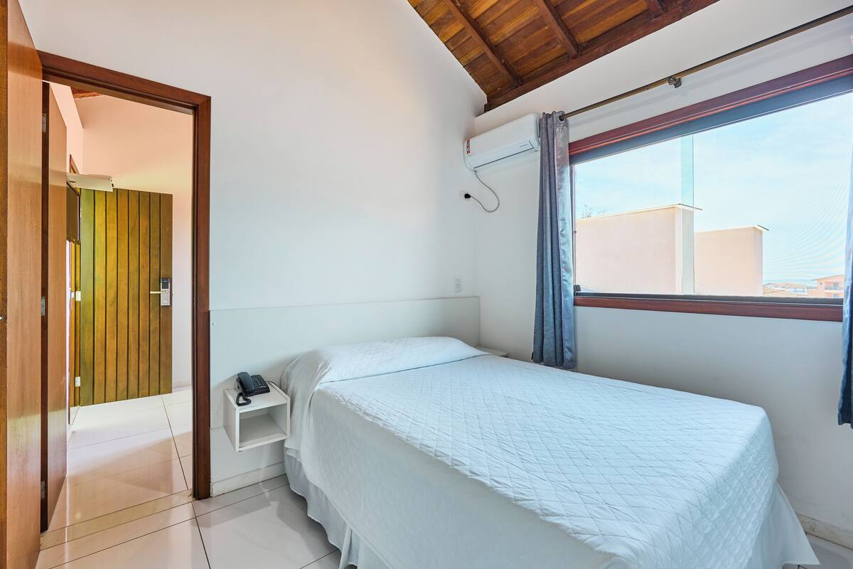 A bedroom with a single bed, a small bedside table with a phone, a window with curtains, and a wooden ceiling.