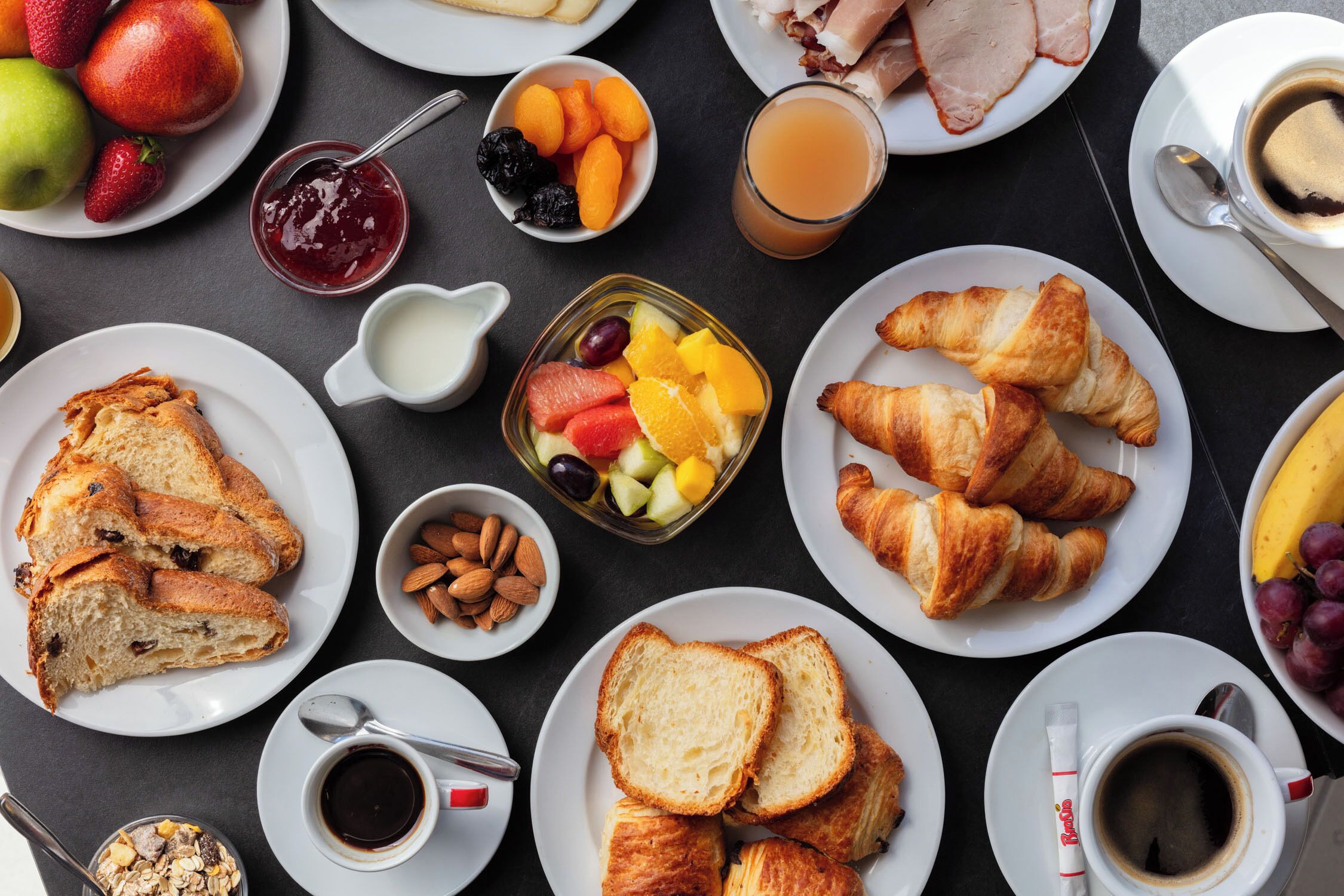 Un assortiment de produits pour le petit-déjeuner, comprenant des croissants, des toasts, des fruits et des boissons, est disposé sur une table.