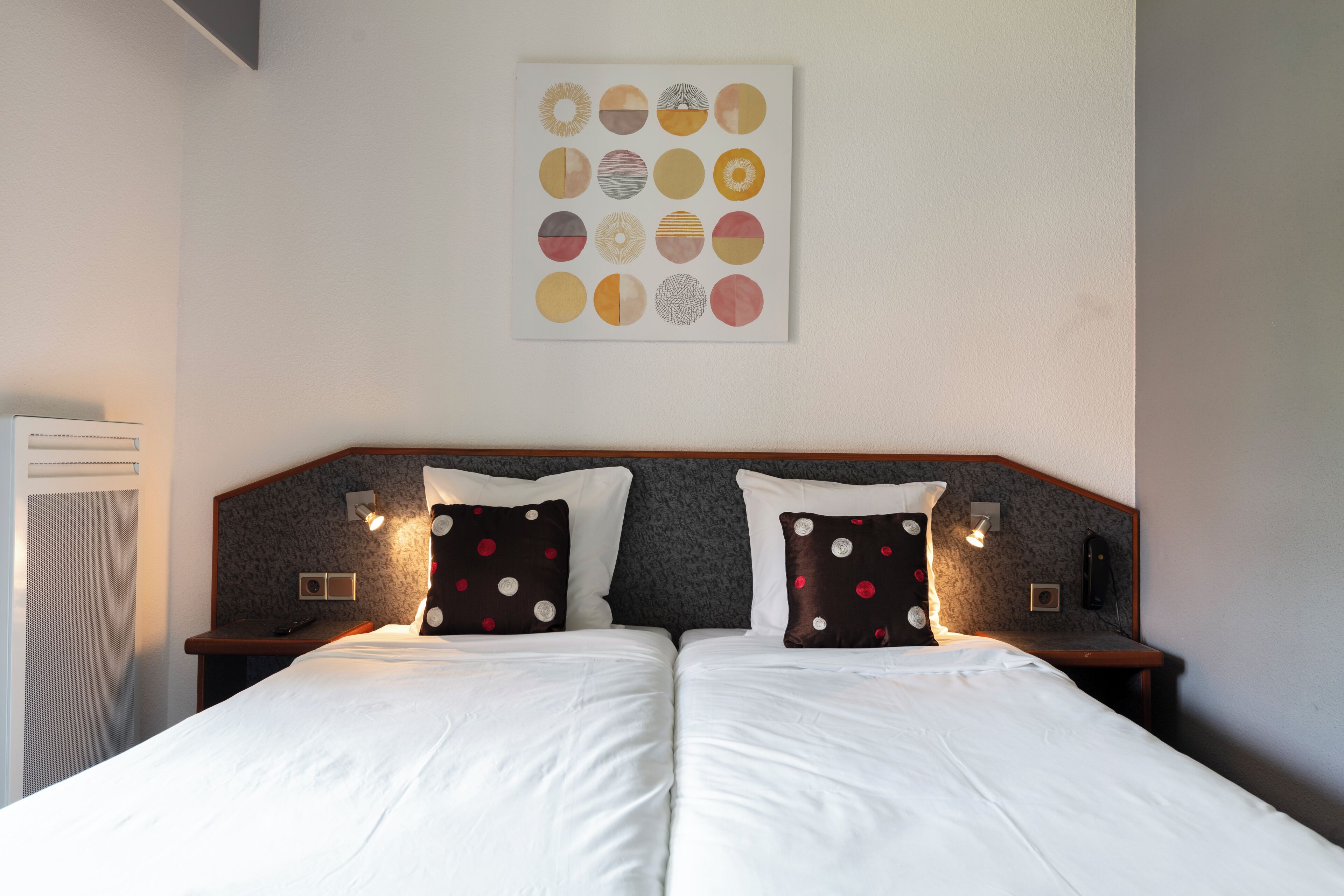A double bed with white linens and two pillows, one with red polka dots and the other with white polka dots, in a room with a headboard and wall-mounted light fixtures.