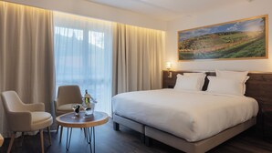 Hotellihuone, jossa on suuri sänky, kaksi tuolia, pieni pöytä, jonka päällä on pullo ja laseja, sekä seinällä oleva maisemamaalaus.