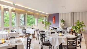 En restaurang med bord uppdukade för servering, stora fönster och krukväxter inomhus.