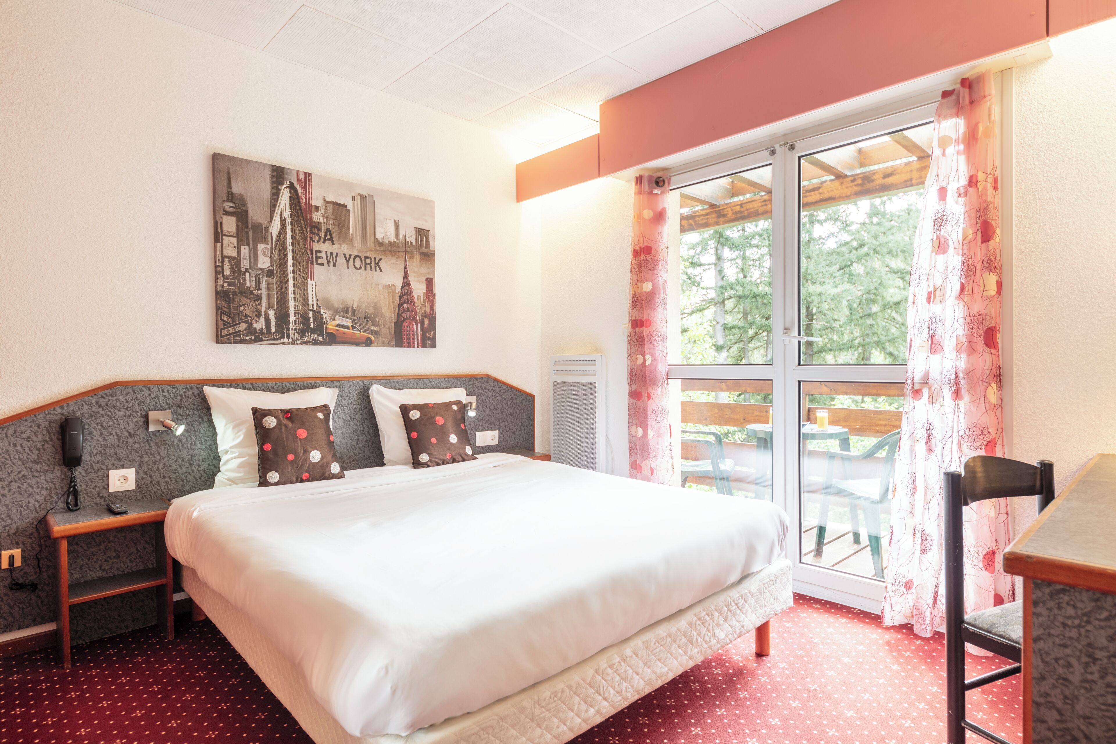 A hotel room with a large bed, a desk, a chair, a window with curtains, and a cityscape painting on the wall.