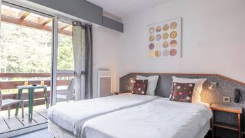 A double bed with white linens, a wooden headboard, and two bedside tables with lamps. A large window with curtains and a view of greenery is visible.