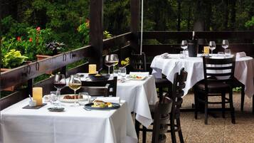 Un área de comedor al aire libre con mesas dispuestas para dos, rodeada de follaje.