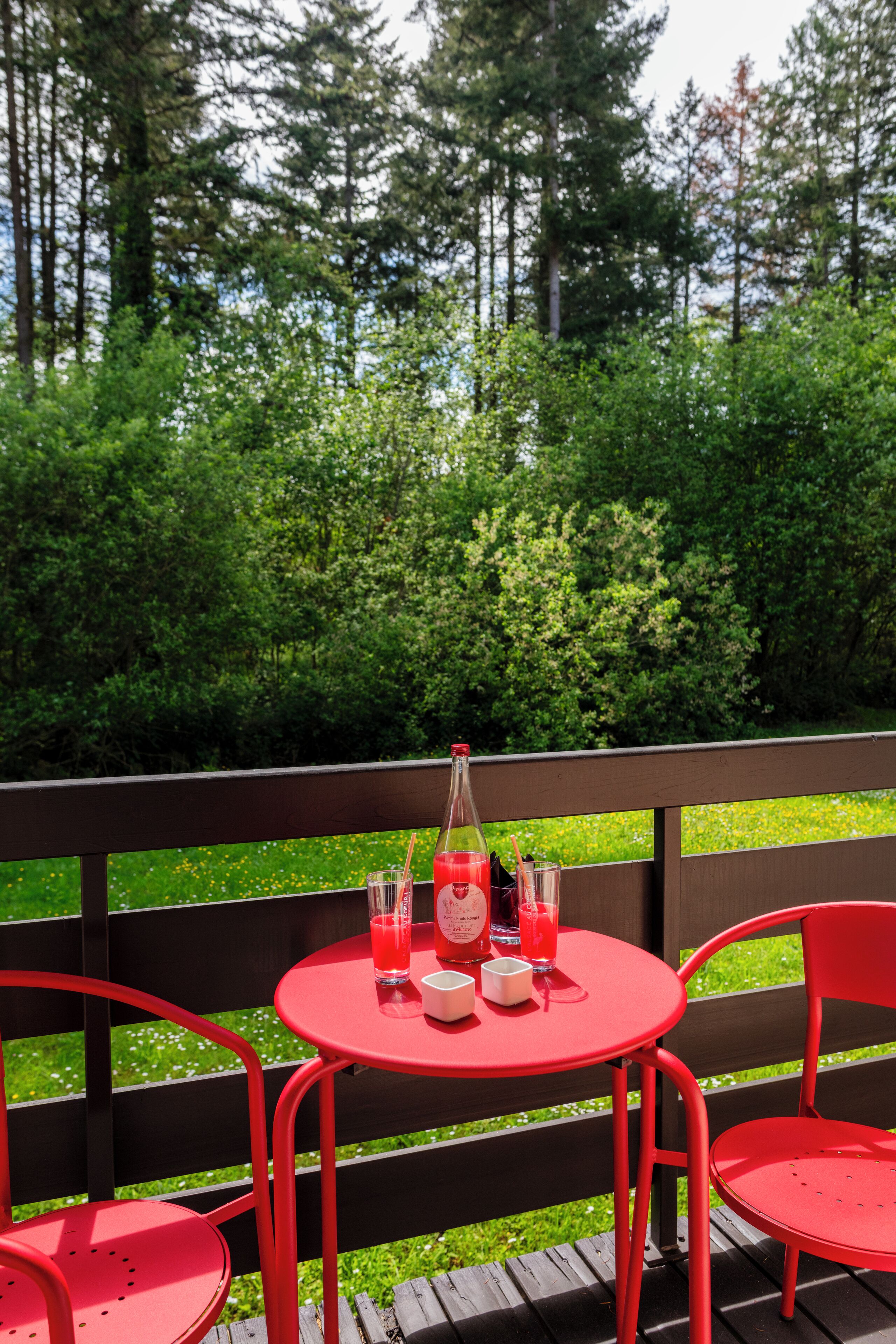 Mesa metálica roja con sillas sobre una terraza de madera, rodeada de vegetación exuberante.
