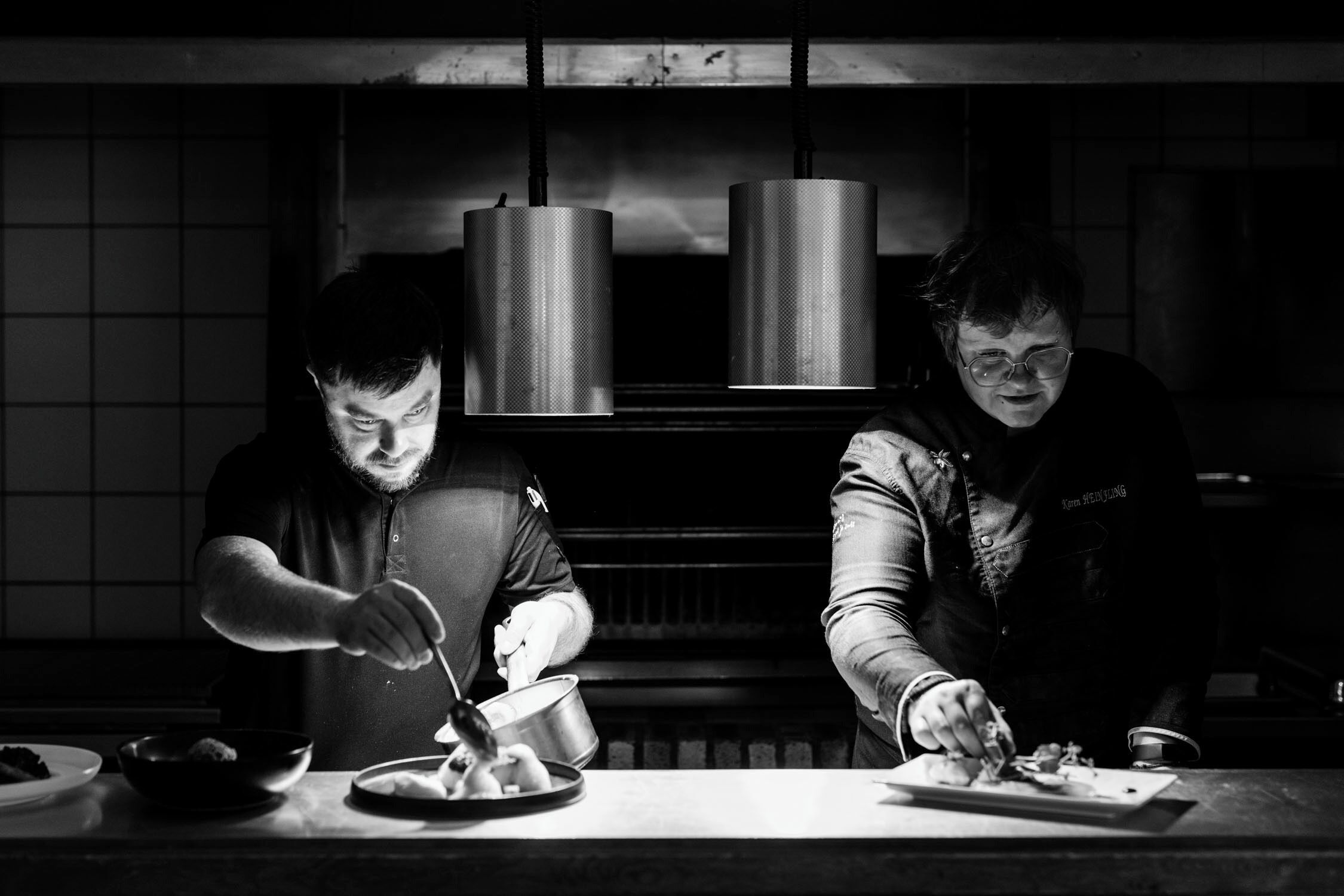 Two chefs preparing food in a kitchen with hanging metal containers above the counter.