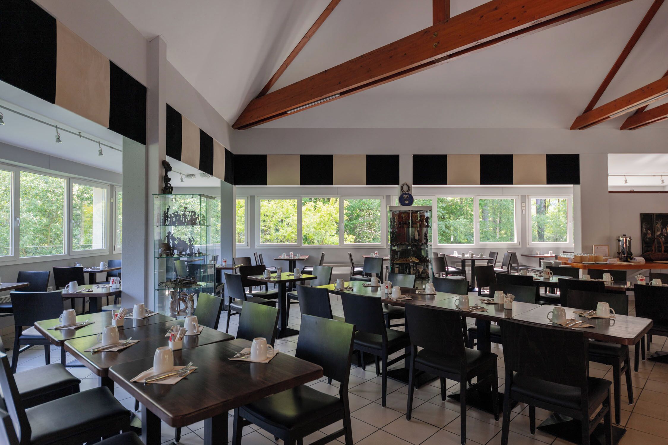 A spacious dining area with multiple tables and chairs, large windows, and a high ceiling with exposed beams.