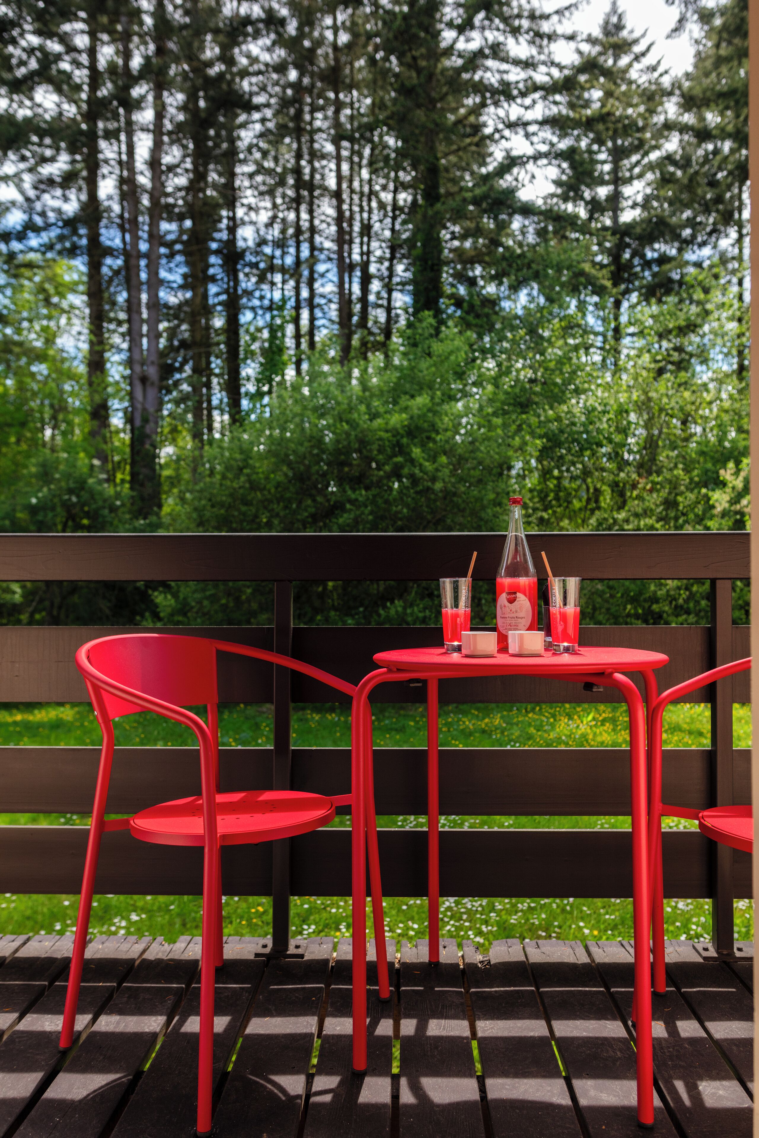 A red metal table and chairs on a wooden deck with a bottle and glasses of red beverage.