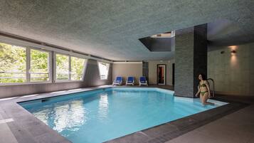 An indoor swimming pool with clear blue water, three blue lounge chairs, and a woman in a green swimsuit.