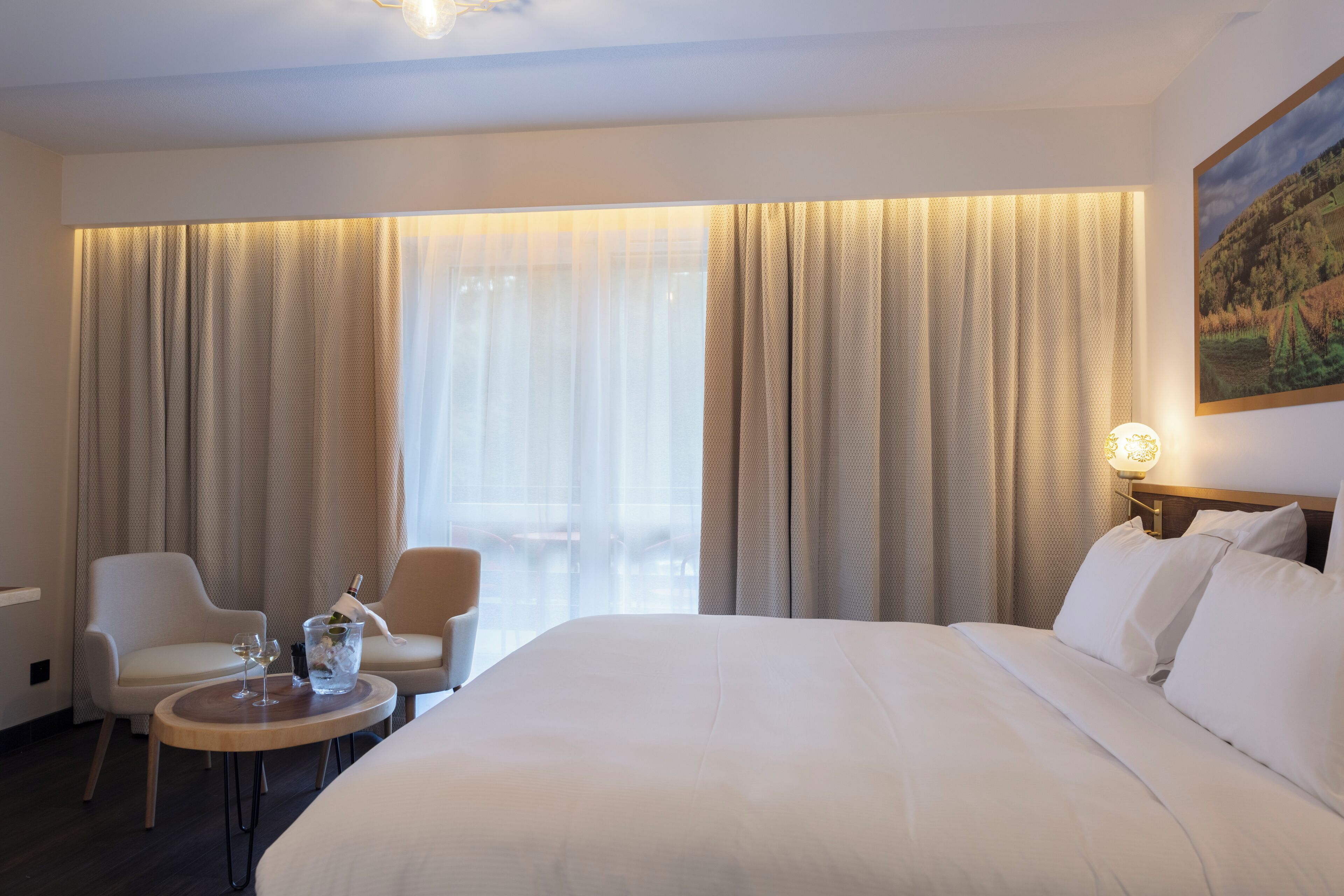 A hotel room with a large bed, two armchairs, a small table with a bottle of champagne and glasses, and a painting on the wall.