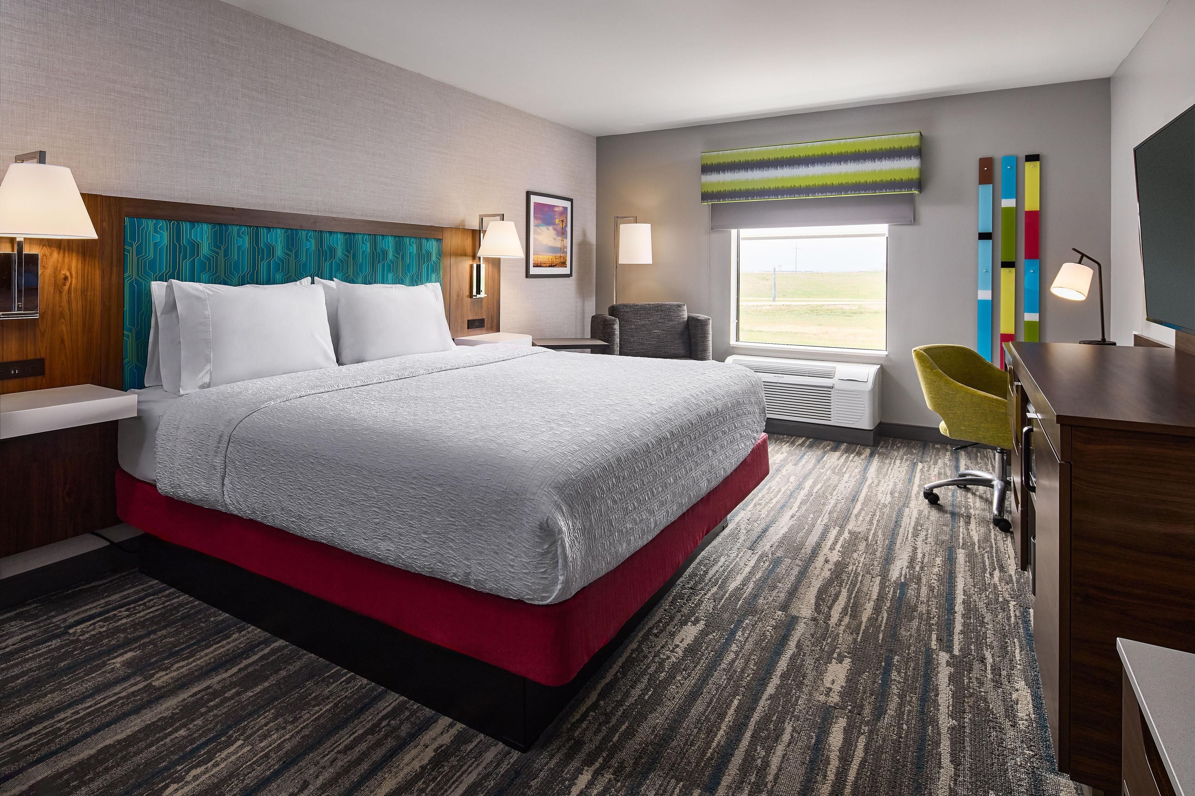 Habitación de hotel con una cama grande, un escritorio, una silla, un ventanal con vistas y un panel de pared colorido.