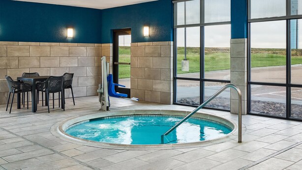 An indoor pool area with a tiled floor, a pool with a metal ladder, a table with chairs, and large windows offering a view of the outdoors. - Hampton Inn Colby (Colby)