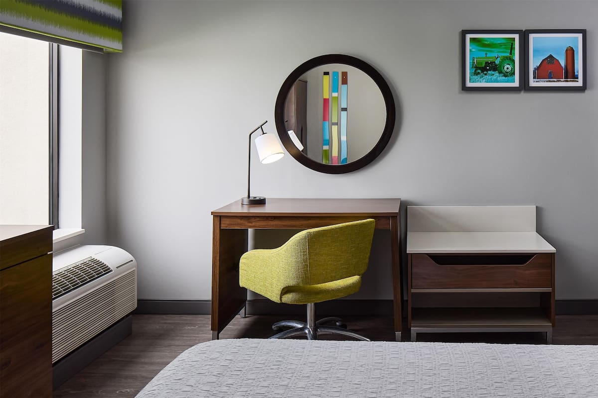 A hotel room with a bed, a desk with a chair, a bedside table, a mirror, and two framed pictures on the wall.