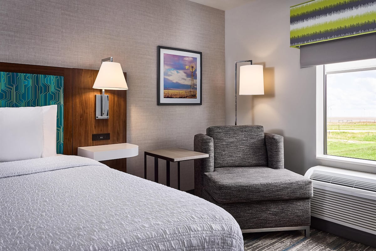 A hotel room with two beds, a grey armchair, a small table, a framed picture on the wall, and a window with a view.