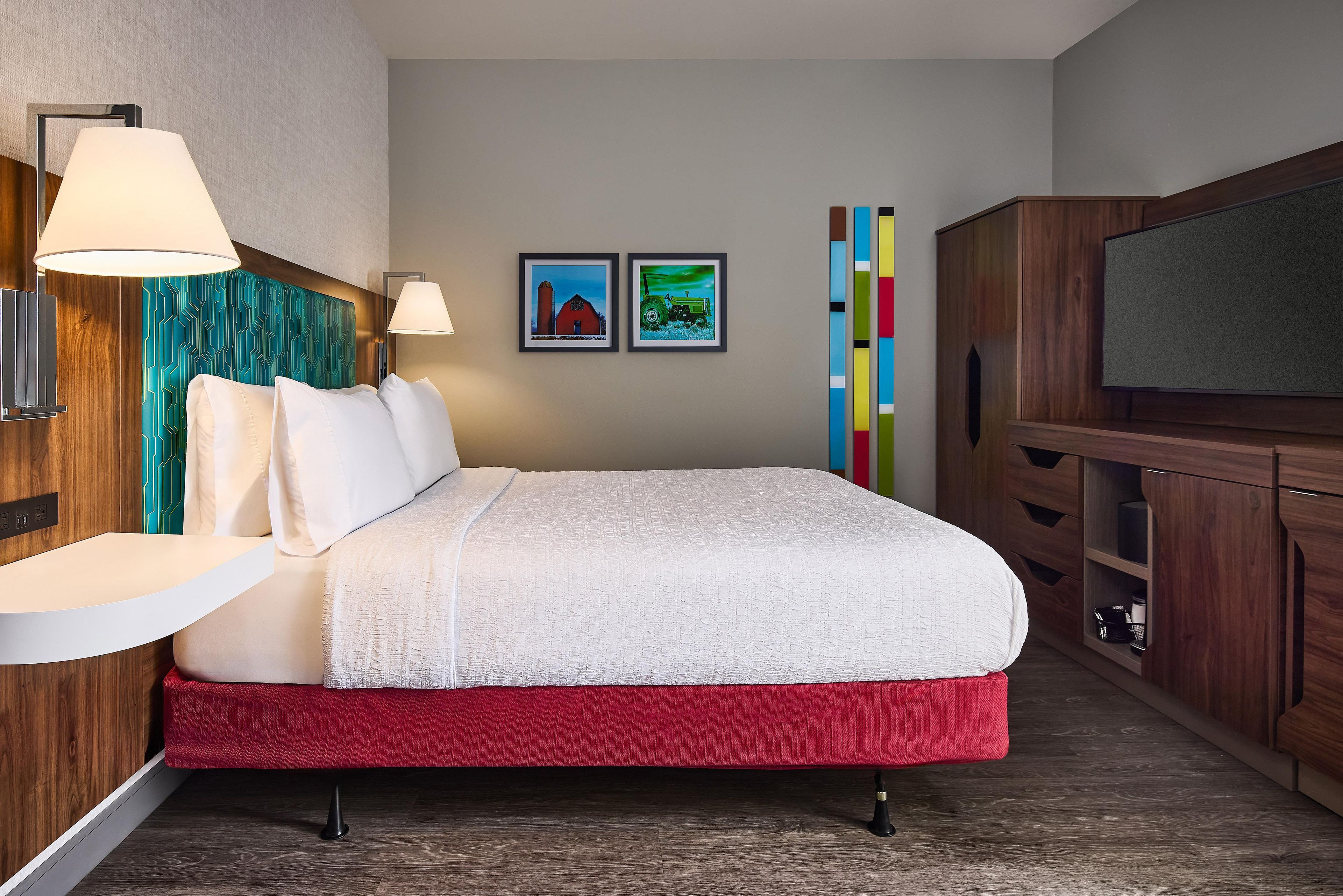 A hotel room with a bed, bedside lamps, a TV, a wooden cabinet, and two framed pictures on the wall.