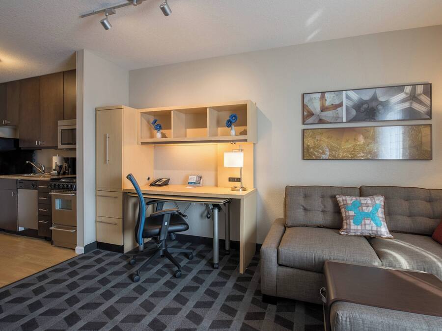 A compact hotel room with a kitchenette, a desk with a chair, a sofa with cushions, and artwork on the walls.