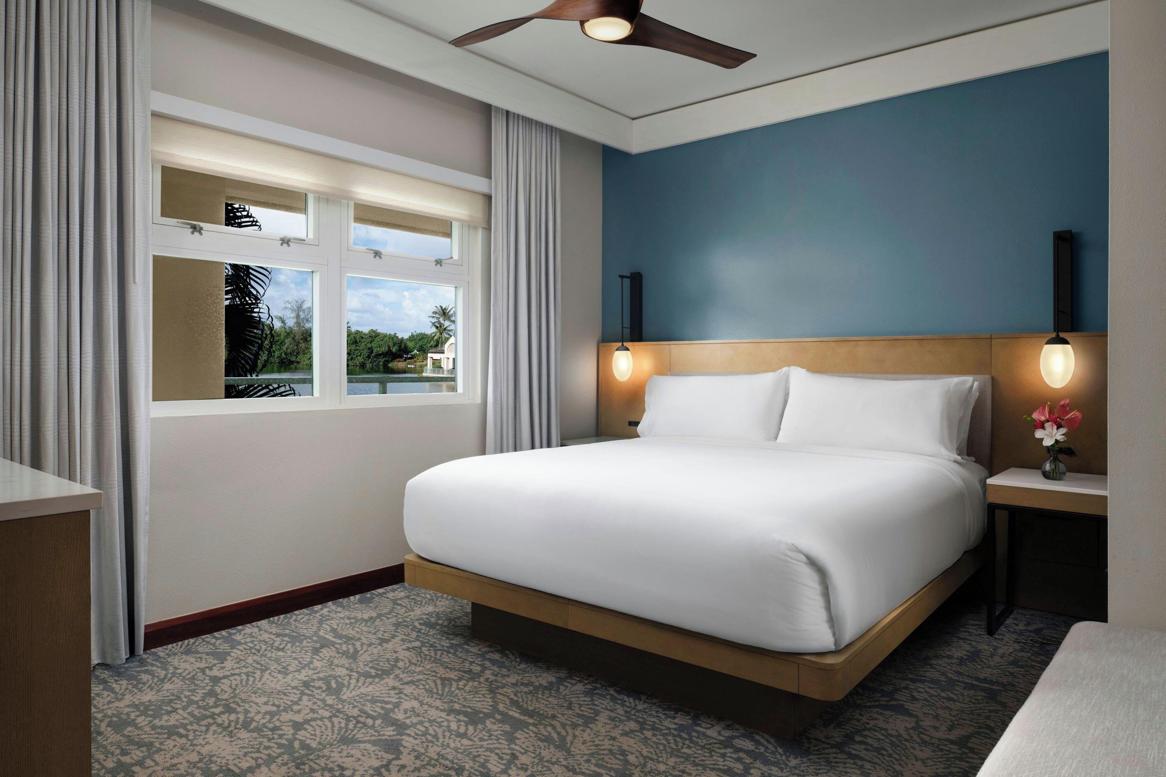A hotel room with a bed, a nightstand, a window with curtains, a ceiling fan, and a view of greenery outside.