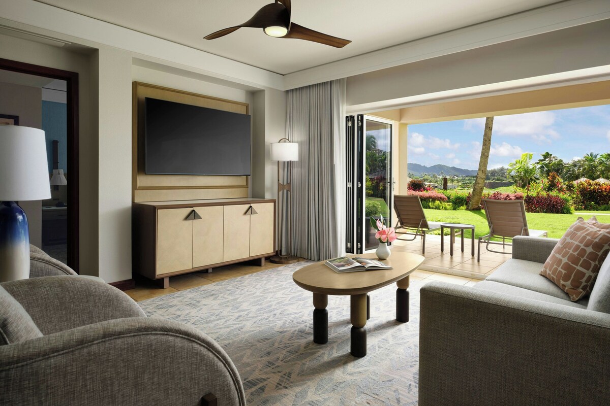 A hotel room with a flat-screen TV, a wooden cabinet, a sofa, a coffee table, and a view of a garden through a sliding glass door.