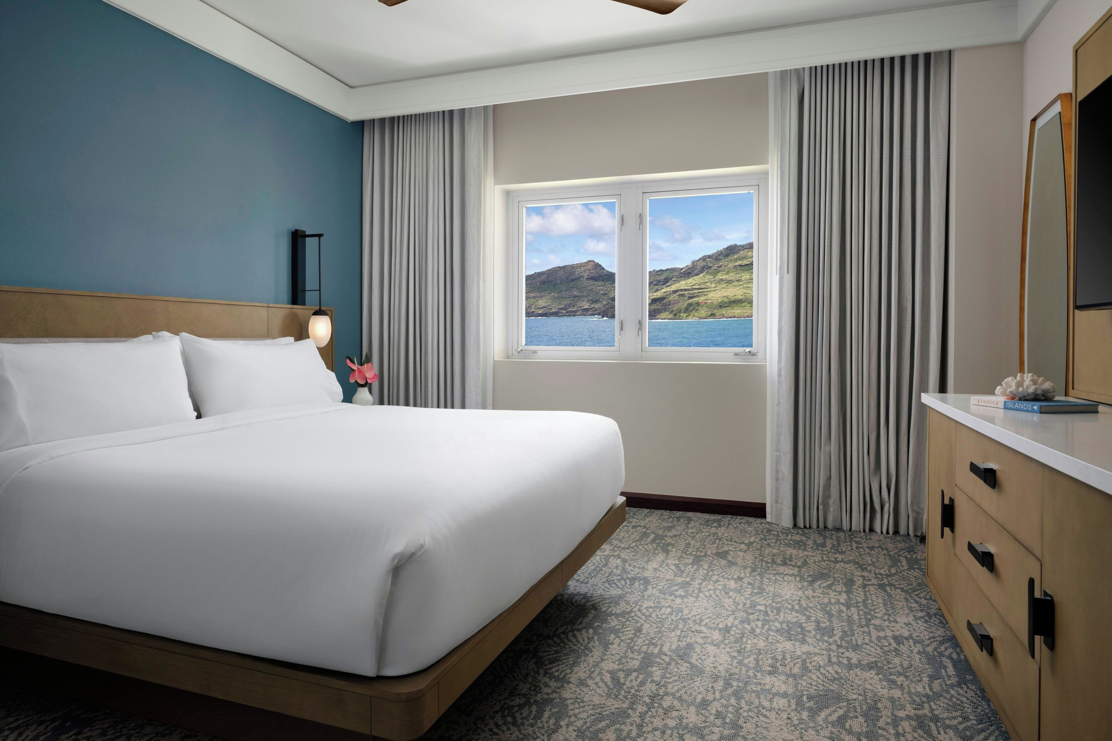 Una habitación de hotel con una cama grande, una ventana que ofrece vistas a colinas y agua, una mesita de noche con una lámpara y un aparador con espejo.