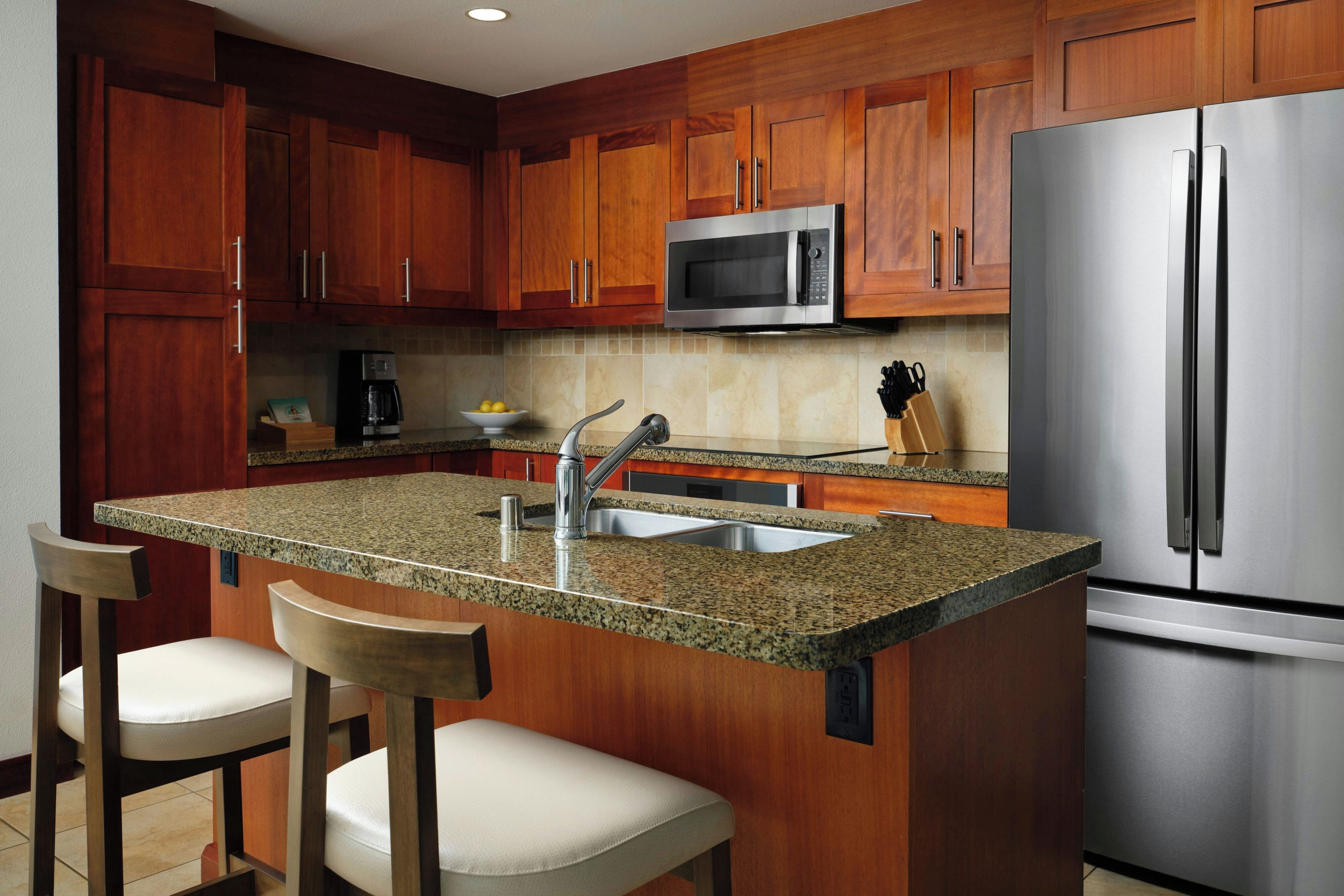 A kitchen with wooden cabinets, a stainless steel refrigerator, a microwave, granite countertops, a sink, and two bar stools.