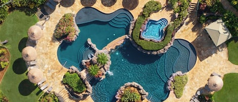 A large outdoor swimming pool with multiple sections and a central island, surrounded by lounge chairs and umbrellas.