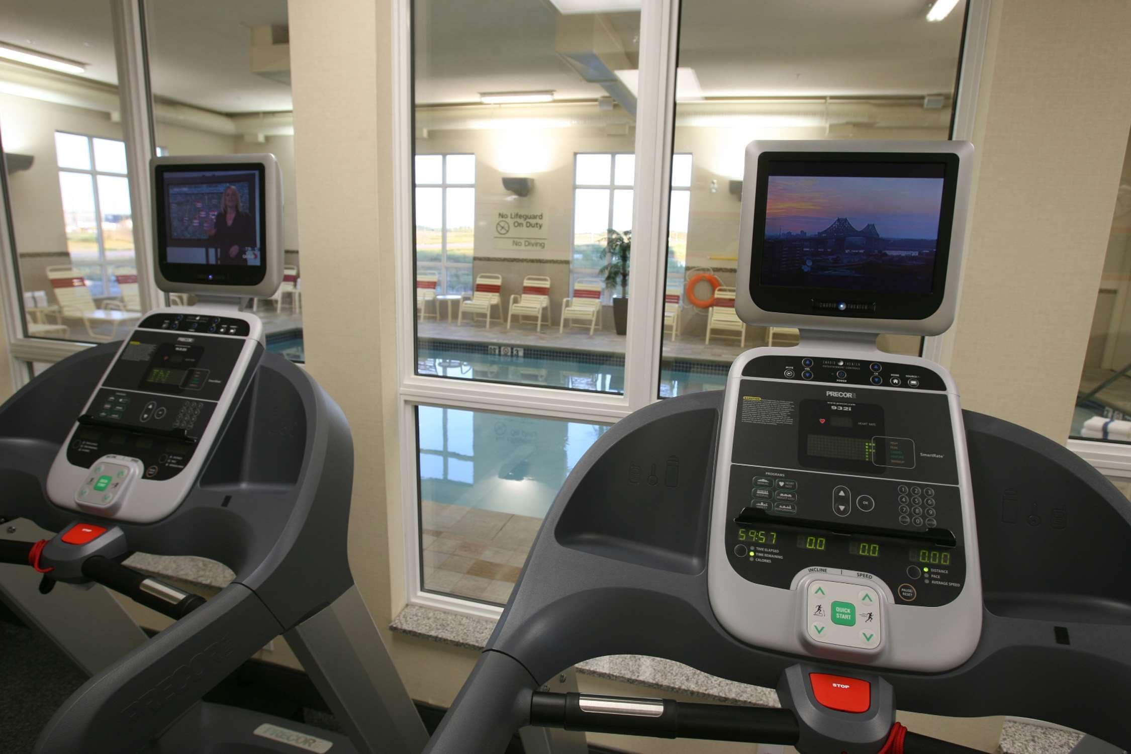 Zwei Trainingsfahrräder mit digitalen Anzeigen, ein durch das Fenster sichtbarer Pool und ein Schild, das darauf hinweist, dass kein Rettungsschwimmer anwesend ist.