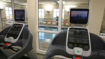 Zwei Trainingsfahrräder mit digitalen Anzeigen, ein durch das Fenster sichtbarer Pool und ein Schild, das darauf hinweist, dass kein Rettungsschwimmer anwesend ist.