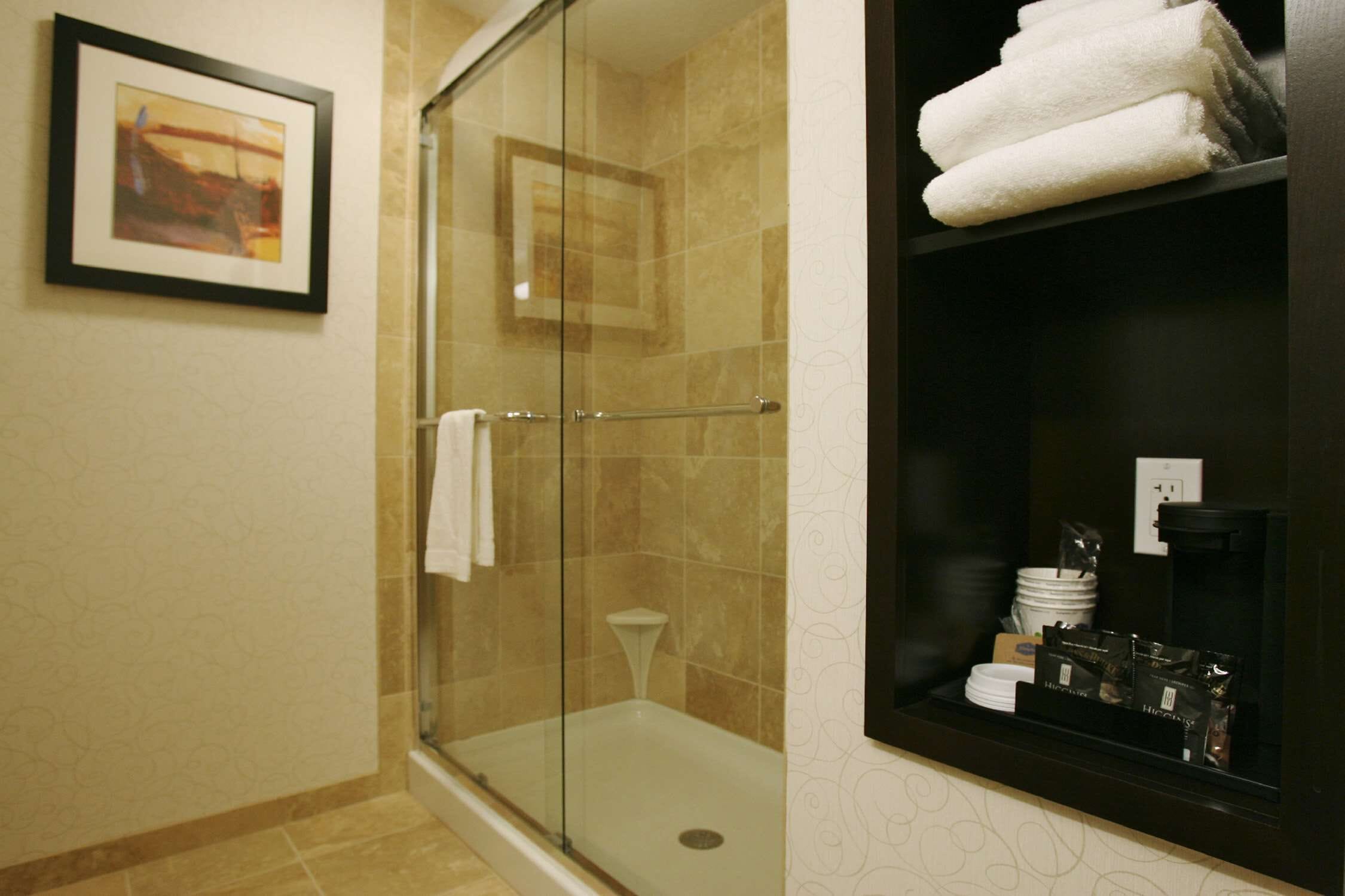 A glass-enclosed shower with a white towel hanging on the door, a framed picture on the wall, and a shelf with towels and toiletries.